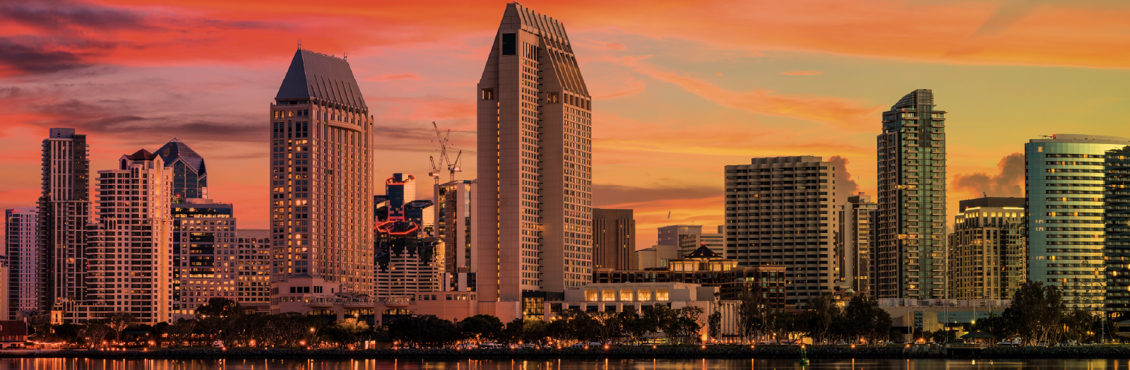 The San Diego skyline at sunset, featuring tall modern buildings reflecting off the calm waterfront. The sky is painted in vivid shades of orange, pink, and yellow, with streaks of clouds adding texture. A gradient border in warm tones frames the top of the image, while a deep blue band anchors the bottom.