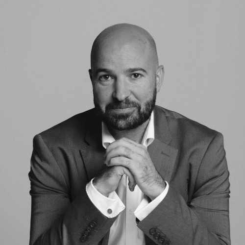 A man with a beard in a suit, resting his hands under his chin and looking at the camera against a plain background.