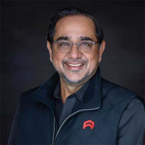Smiling man with glasses and short dark hair wearing a black quilted vest with a red logo in a studio headshot against a dark background.