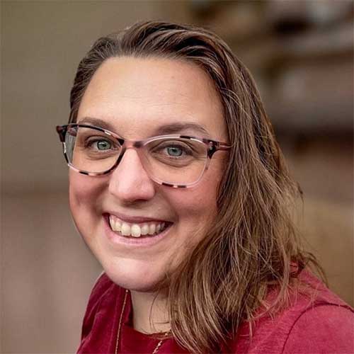 Smiling woman with shoulder-length brown hair and rectangular glasses in a close-up headshot with a softly blurred background.