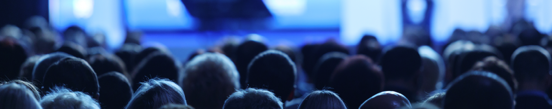 Audience seated facing a stage at a conference, seen from behind under blue lighting.