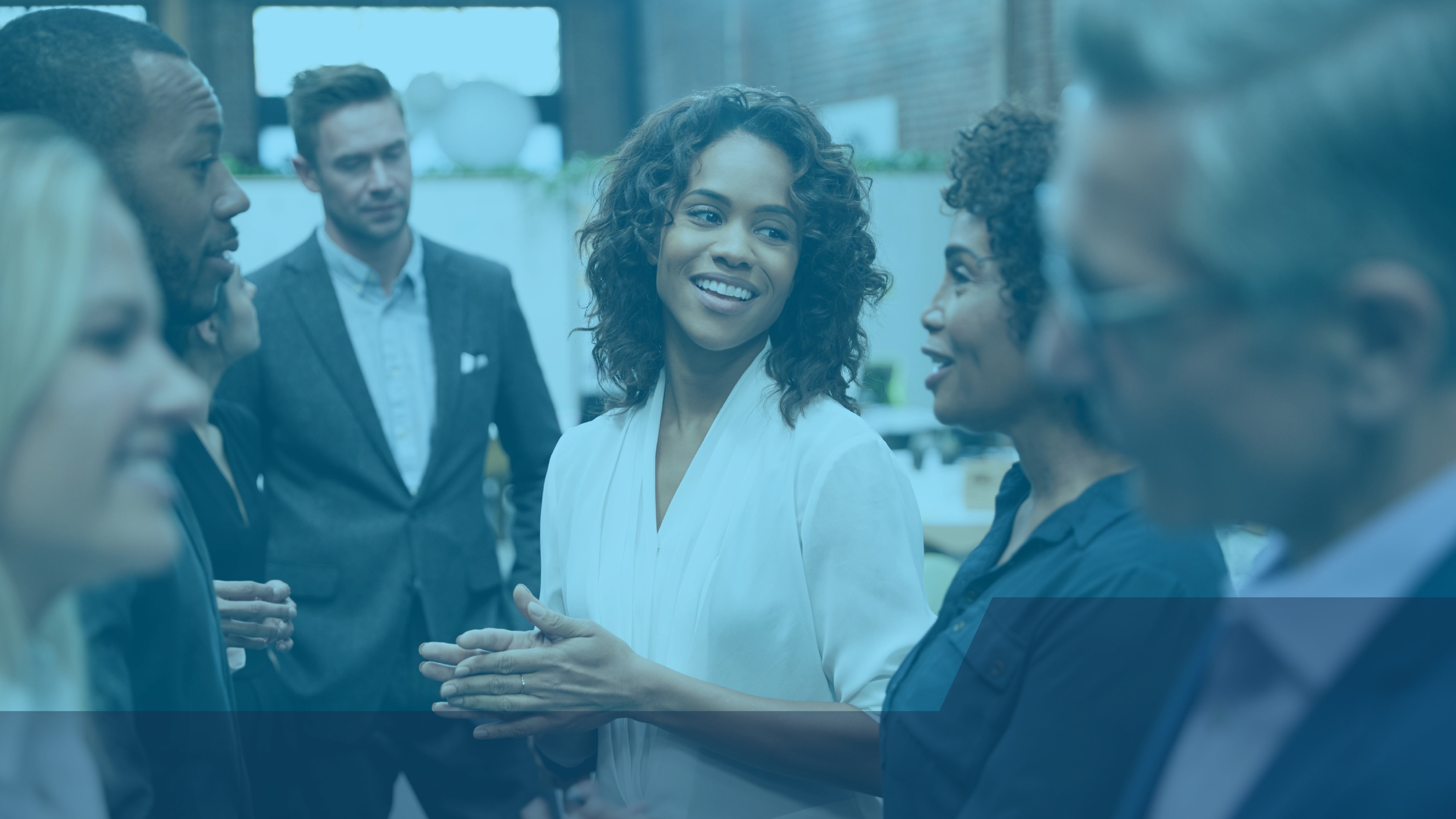 A group of professionals standing together and conversing, with a woman in a white blouse smiling at a colleague in the center of the group.