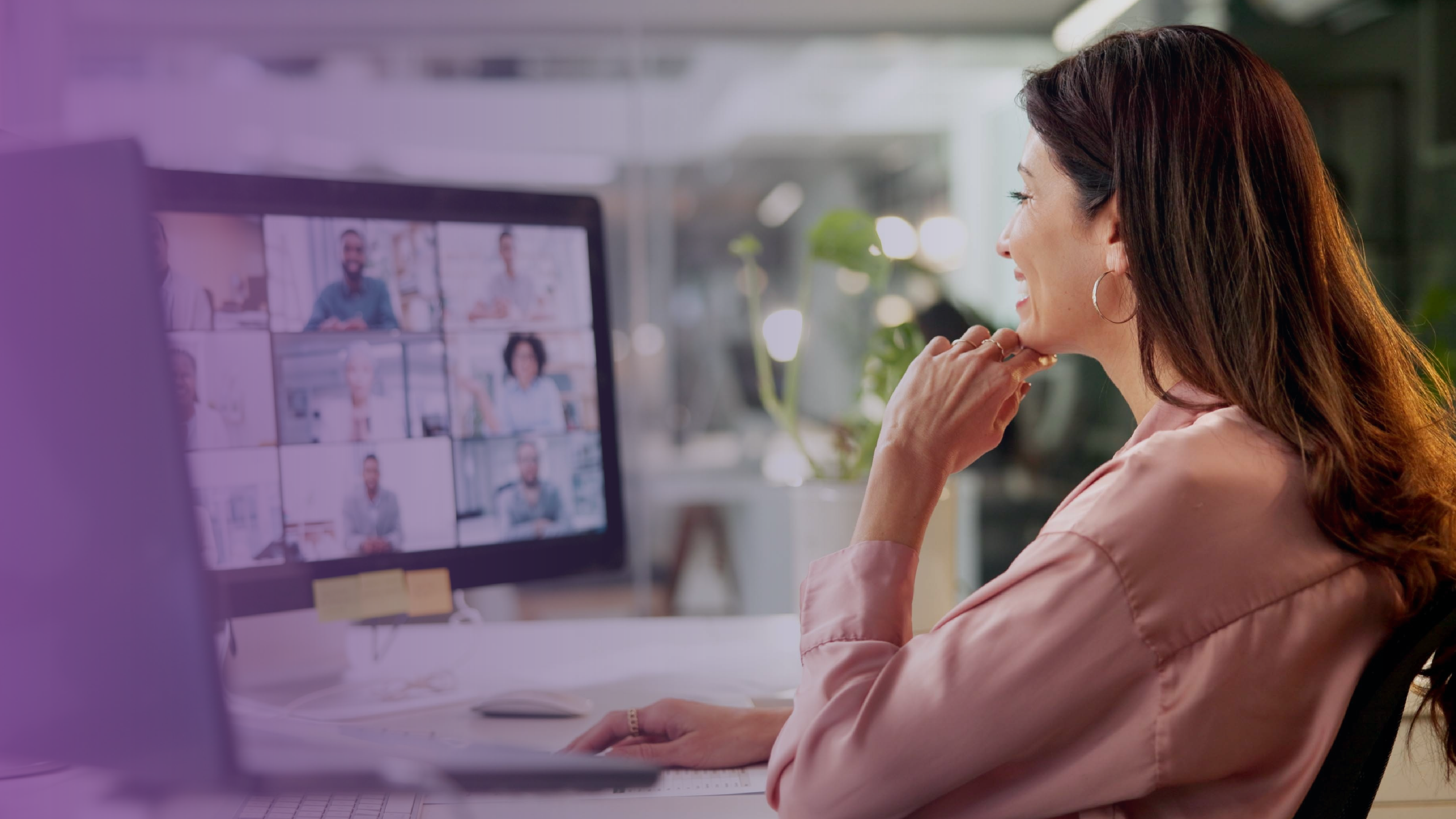 Woman in a pink blouse sitting at a desk and smiling while participating in a virtual meeting on her computer, with multiple people visible on the screen.